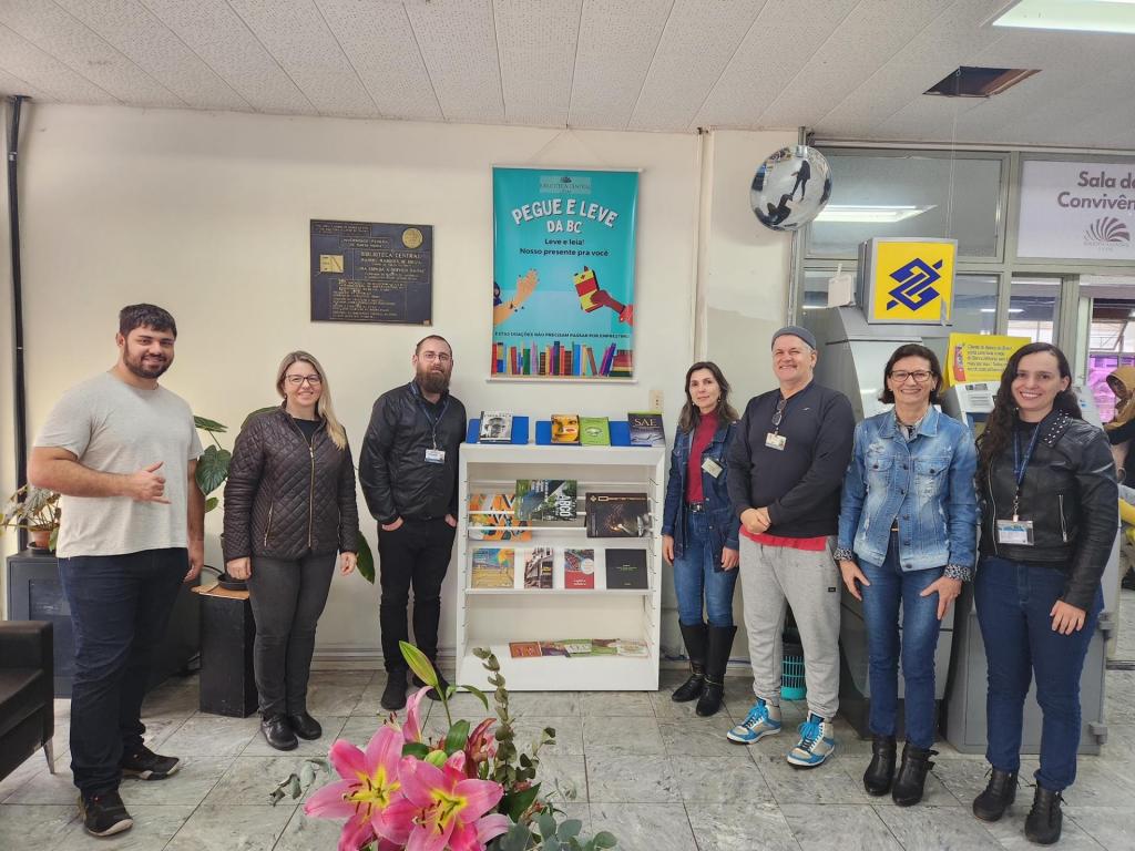 Pessoas reunidas ao redor de uma estante de metal baixa, conde há livros expostos e livros embalados como presente. Na parede atrás da estante está pendurado um banner onde está escrito "Pegue e leve da BC".