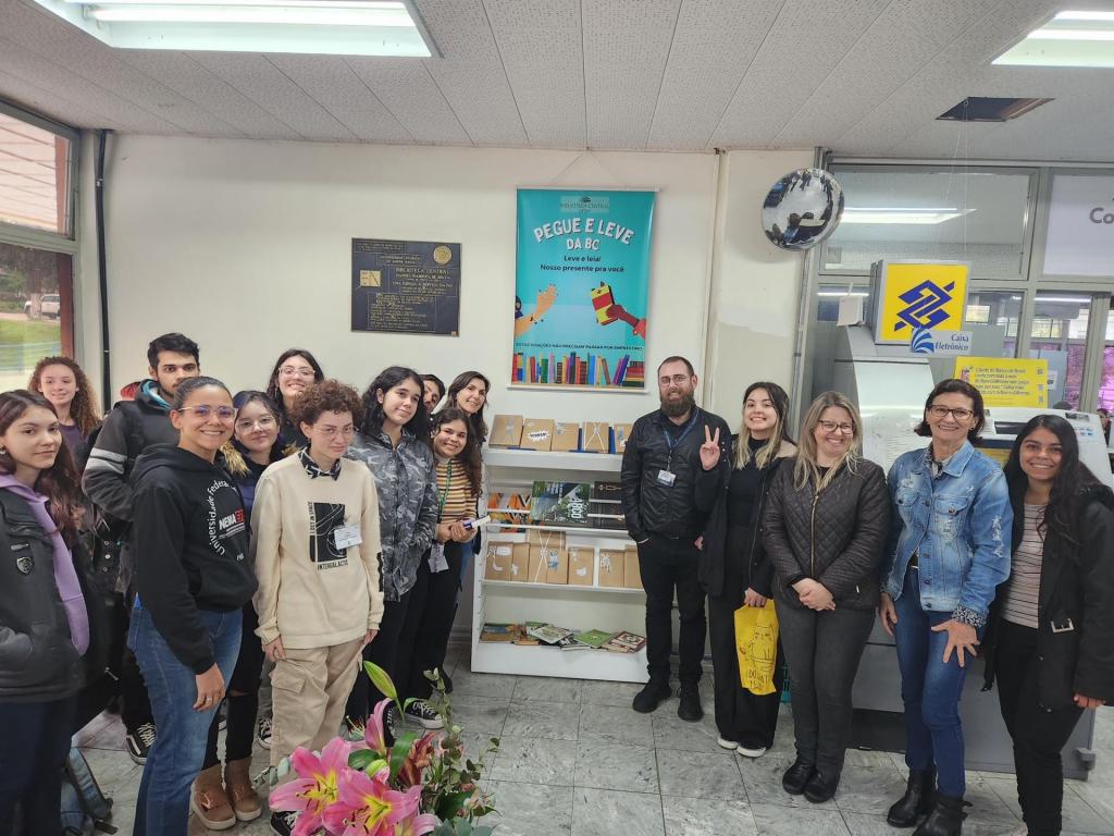 Pessoas reunidas ao redor de uma estante de metal baixa, conde há livros expostos e livros embalados como presente. Na parede atrás da estante está pendurado um banner onde está escrito "Pegue e leve da BC".