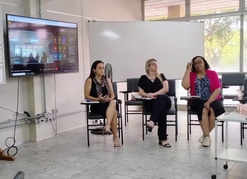 Foto de três mulheres sentadas (Lizandra, Paula e Deisiré) ao lado de um monitor exibindo a transmissão online da reunião para pessoas de forma remota. As três mulheres olham para a frente onde está o público.