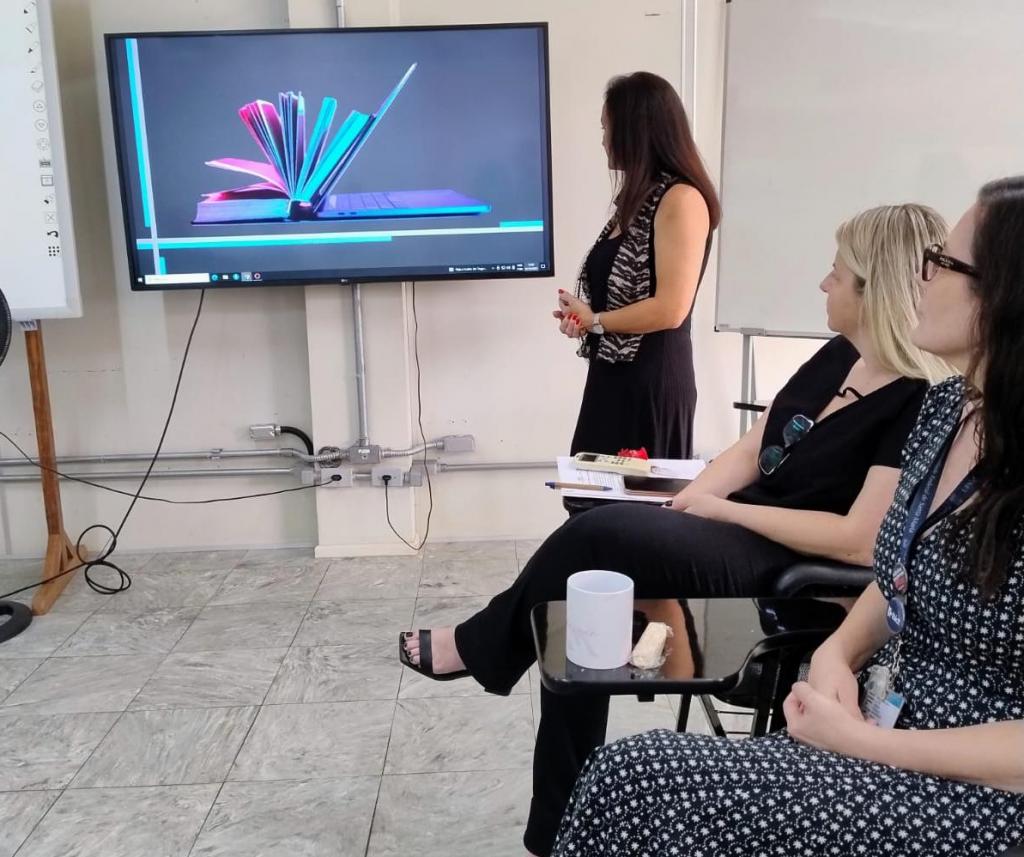 Foto de 3 pessoas enquadradas e de um monitor exibindo a projeção de uma apresentação de slides. Uma mulher branca (Lizandra) está em pé olhando para a apresentação, e outras duas sentadas assistindo.
