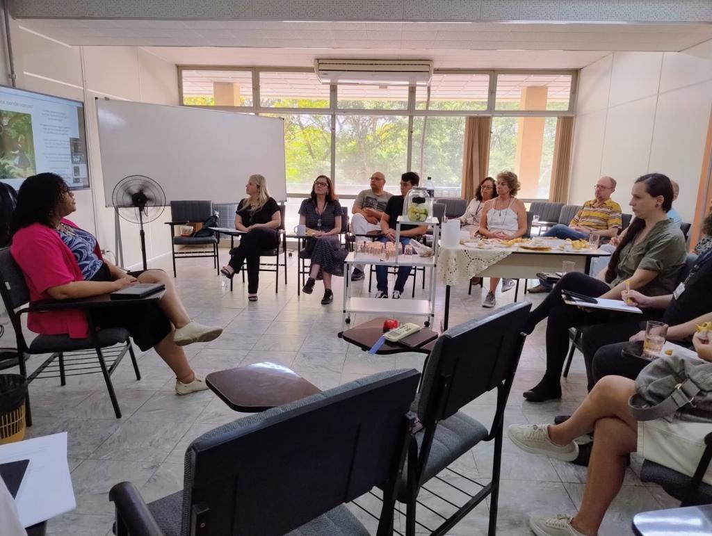Foto de um grupo de mais de 9 pessoas enquadradas, além de outras pessoas presencialmente e um monitor exibindo a transmissão online da reunião para pessoas de forma remota. À esquerda uma mulher negra (Deisiré) está sentada à frente do grupo. Também é visível uma mesa de salgadinhos e bebidas geladas.