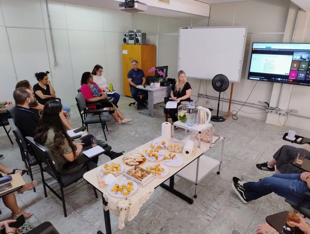 Foto panorâmica de um grupo de mais de 8 pessoas enquadradas, além de outras pessoas presencialmente e um monitor exibindo a transmissão online da reunião para pessoas de forma remota. No centro há uma mesa de salgadinhos. Uma mulher branca (Paula) está sentada à frente no centro do grupo.