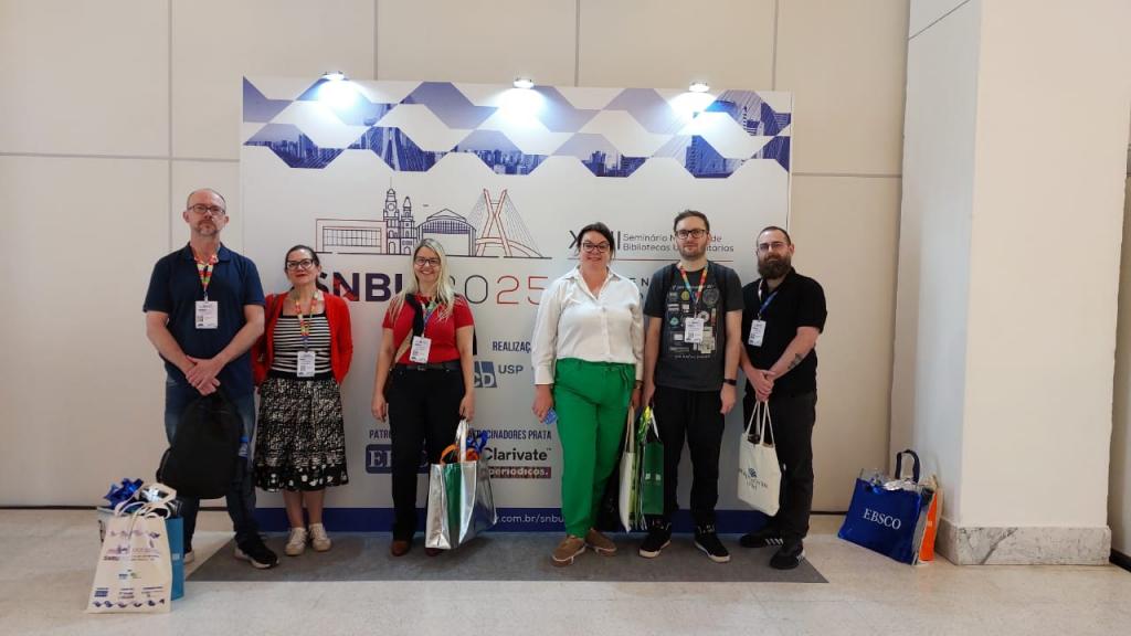 Foto de seis pessoas posando em frente a um backdrop do 23. SNBU. As pessoas, três homens e três mulheres, todos brancos, estão identificadas com os crachás do evento e algumas delas seguram sacolas nas mãos.