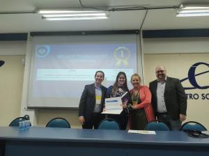 Foto retangular mostrando duas mulheres e dois homens em frente a uma tela projetada com o trabalho vencedor do evento