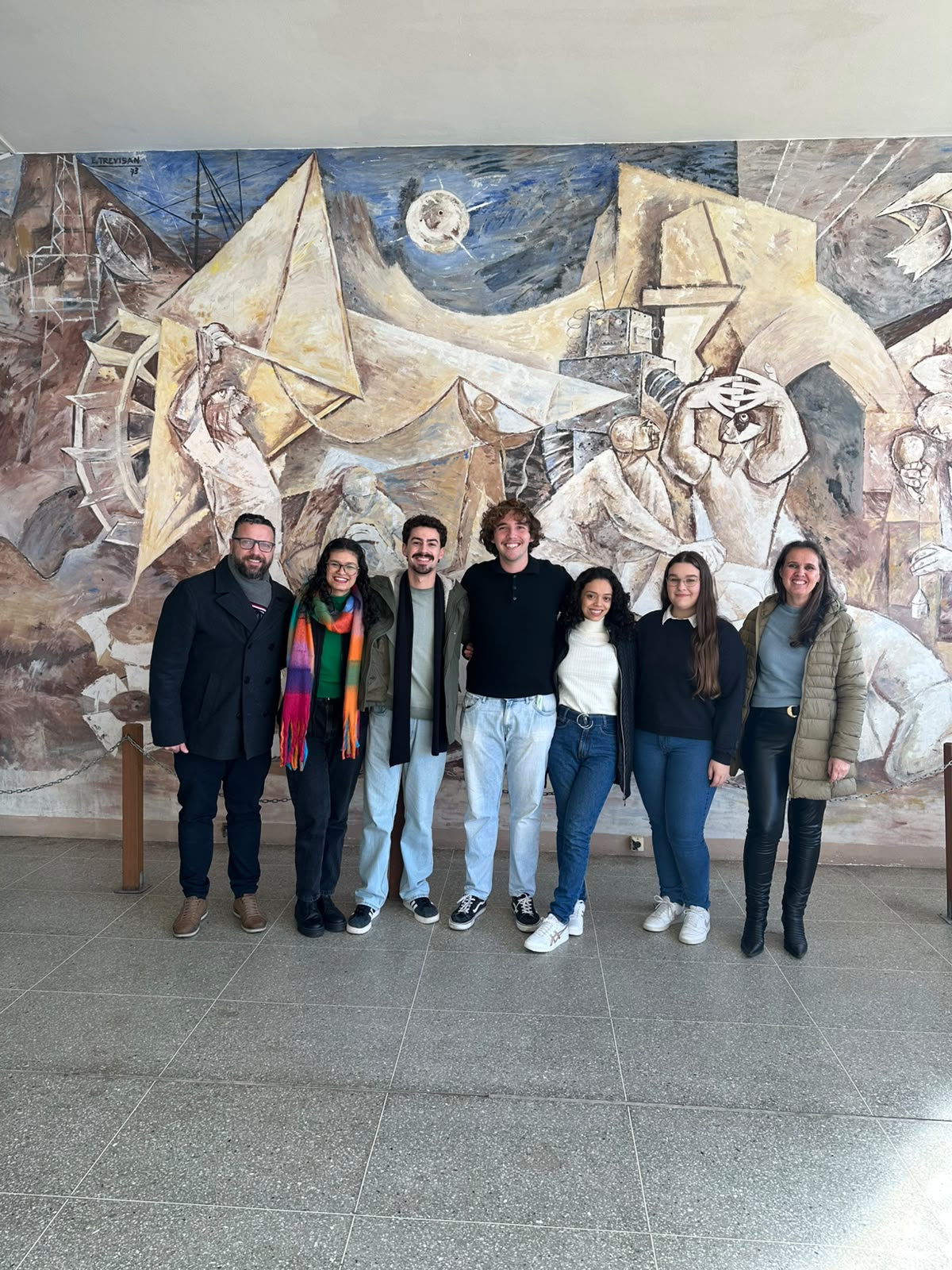 Sete pessoas posam lado a lado em um saguão do Centro de Tecnologia da UFSM, em frente a um grande mural artístico. Na foto estão o professor Elvis, cinco estudantes selecionados para o Programa BRAFITEC — três mulheres e dois homens — e a vice-diretora do CT. Todos sorriem para a câmera e estão em pé, lado a lado. O mural ao fundo apresenta formas geométricas e elementos relacionados à ciência e à tecnologia.