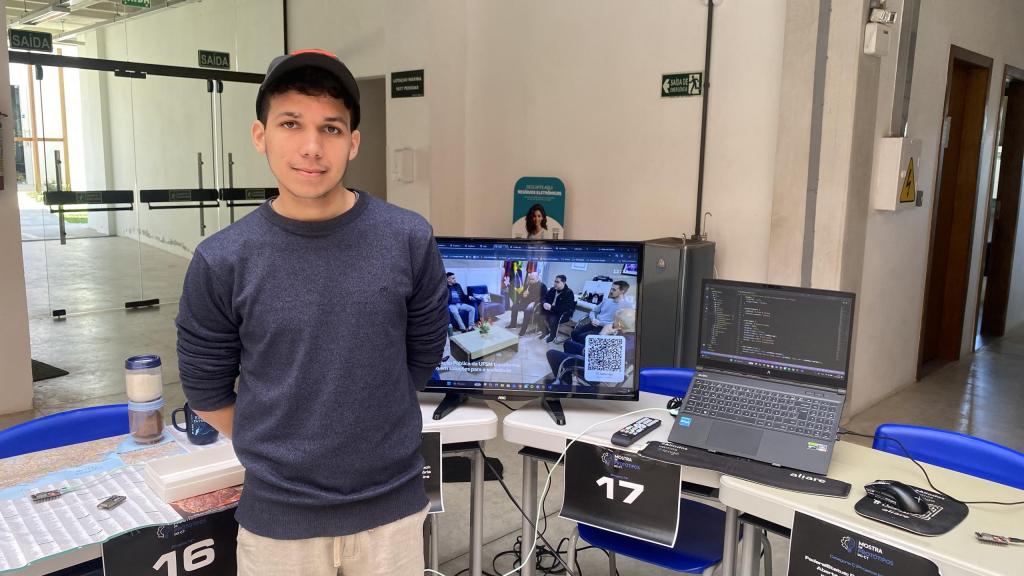 Fernando Dorneles, jovem com boné preto e blusa de moletom azul escura, em pé atrás de uma mesa de exposição na Mostra de Protótipos. Ele olha para a câmera e sorri. À sua frente estão monitores exibindo a plataforma Academic Companion e códigos de programação, identificados com placas "16" e "17". O fundo mostra portas de saída de emergência com placas "Saída".