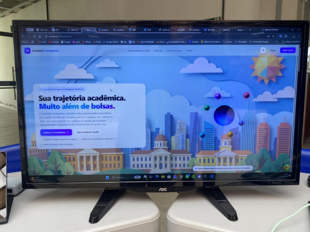 Monitor de computador marca AOC em uma mesa de exposição exibindo a tela inicial do site Academic Companion. A página tem fundo azul com ilustrações coloridas de prédios e céu com nuvens, destacando em letras grandes o título "Sua trajetória acadêmica. Muito além de bolsas" e botões de navegação.