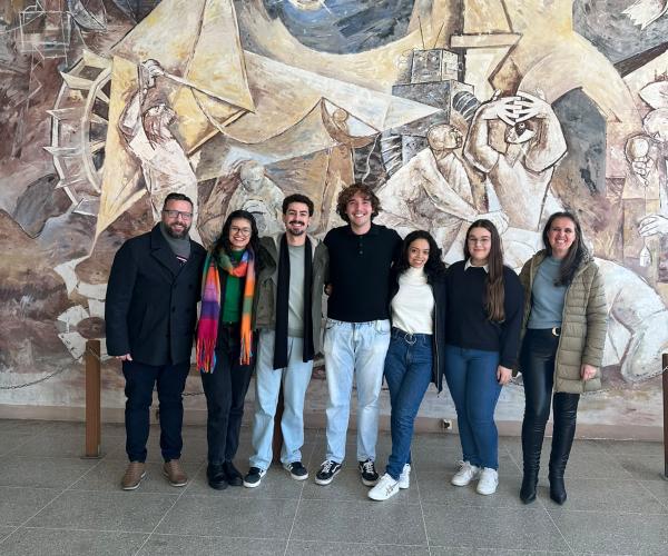 Sete pessoas posam lado a lado em um saguão do Centro de Tecnologia da UFSM, em frente a um grande mural artístico. Na foto estão o professor Elvis, cinco estudantes selecionados para o Programa BRAFITEC — três mulheres e dois homens — e a vice-diretora do CT. Todos sorriem para a câmera e estão em pé, lado a lado. O mural ao fundo apresenta formas geométricas e elementos relacionados à ciência e à tecnologia.