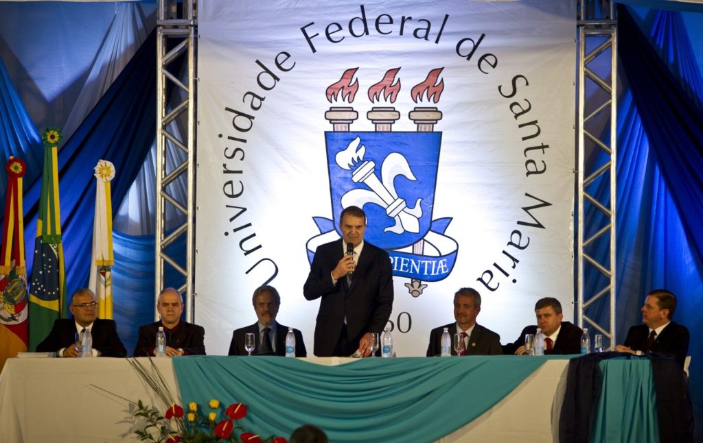 Uma mesa com sete homens. Ao fundo, uma banner com a logo da UFSM. O homem no centro da mesa está em pé, falando com um microfone em sua mão direita.