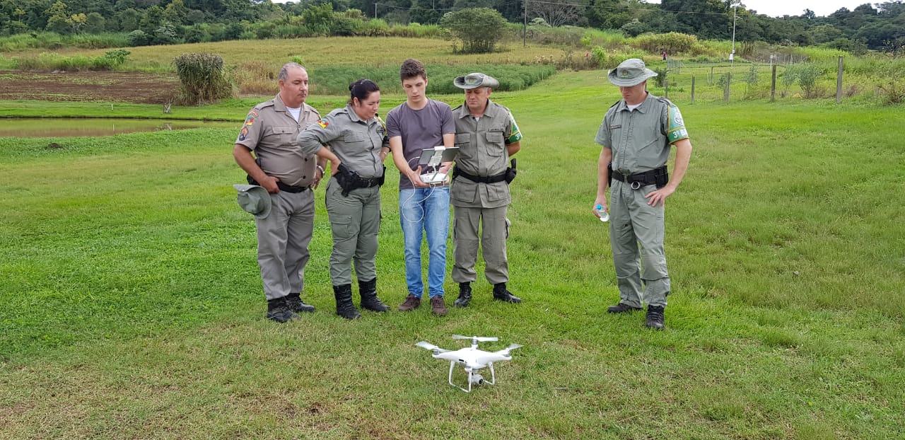 Quatro patrulheiros ambientais com farda cinza, olham o tablet que monitora o drone. O tablet é segurado por um aluno branco de cabelos morenos, que está vestindo calça jeans e camiseta roxa. O drone está no chão, em frente a todos eles.