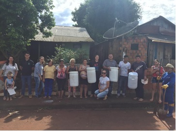 projeto reciclagem de resíduos sólidos. Cerca de 10 pessoas, uma ao lado da outra, em frente a duas casas, recebem quatro bombonas brancas que são usadas para compostagem doméstica