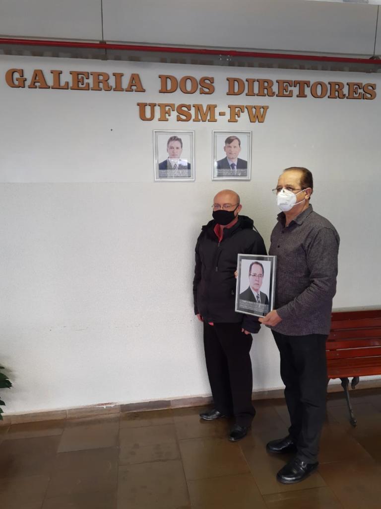 Na imagem, da esquerda para a direita, aparecem o professor Bráulio Caron, atual diretor da UFSM-FW, e o professor Lauro Chielle, primeiro diretor Pro Tempore da UFSM-FW, segurando a foto do professor Lauro. A imagem foi registrada em frente à Galeria de Diretores da UFSM-FW.