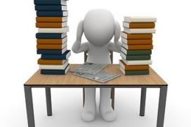 Descrição da imagem: Boneco na cor branca sentado em uma cadeira de madeira em frente a uma mesa de madeira. Em cima da mesa estão duas pilhas de livros, sendo a da esquerda mais alta que a da direita. Entre as pilhas de livro estão duas folhas de papel. O boneco está com as mãos na cabeça.