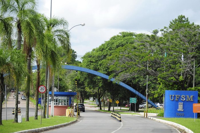 Descrição da imagem: Fotografia do pórtico de entrada da Universidade Federal de Santa Maria, destacando o arco na cor azul e o brasão da UFSM fixado no muro lateral à direita. Árvores também compõem a imagem.