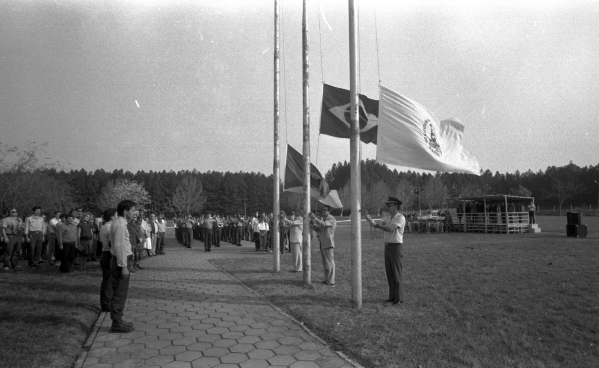 Foto preto e branca de pessoas em posição de respeito às bandeiras flamulantes no largo do Planetário