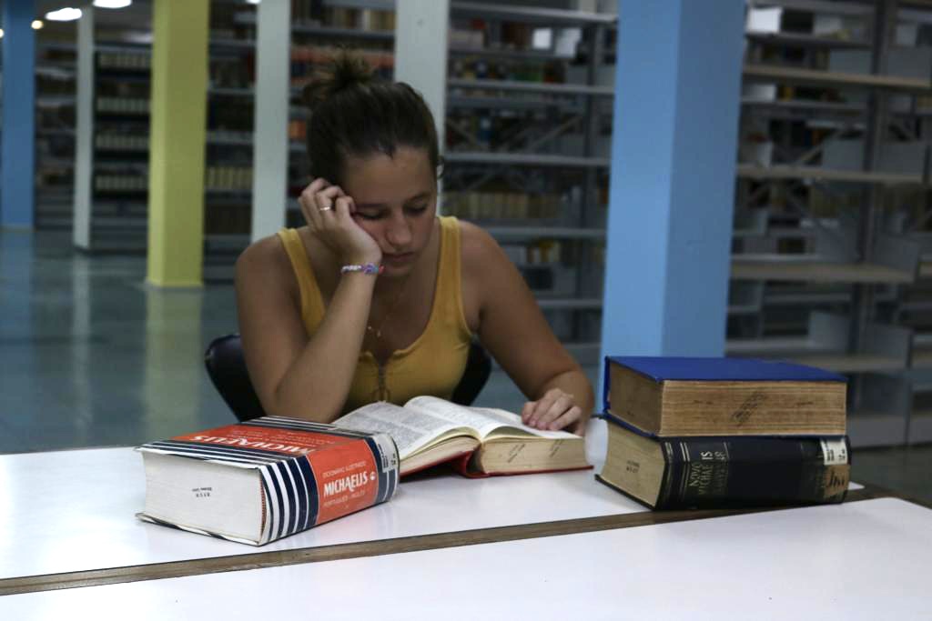 A imagem é a fotografia de uma estudante branca, que possui cabelo castanho claro, amarrado em um coque, e usa uma regata amarela. Ela está sentada em uma mesa da biblioteca, e tem no rosto uma expressão de cansaço, enquanto lê um livro. Sua cabeça está apoiada na mão direita, como se estivesse tentando se manter acordada.