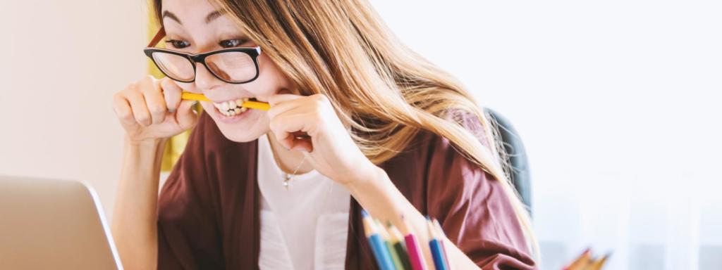 Mulher asiática usando óculos morde lápis ao olhar para tela do computador.