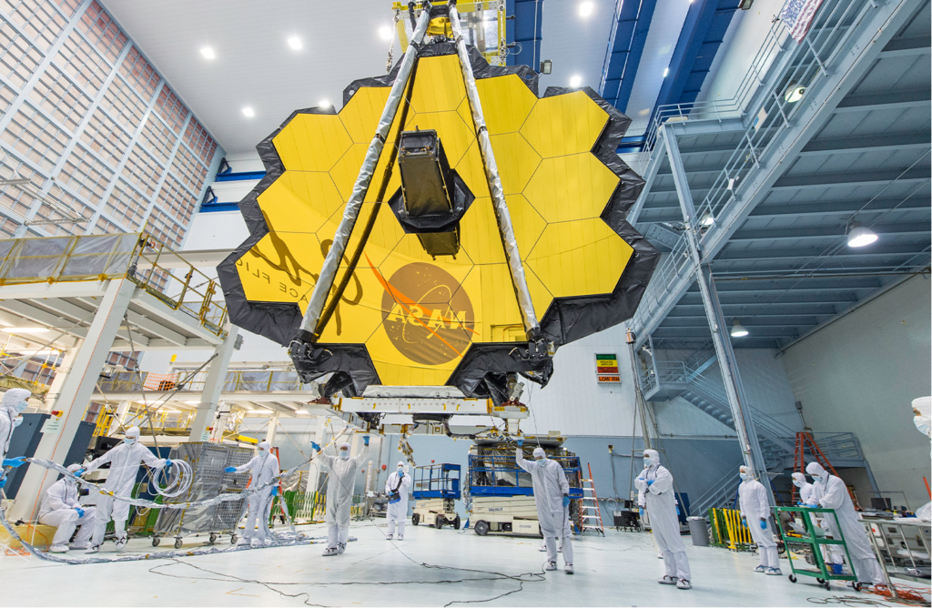 Fotografia horizontal e colorida de um telescópio amarelo e preto. No centro da imagem, o telescópio, em tamanho muito grande e no formato de uma colmeia de abelhas, e amarelo. No centro, a estrutura do cano de observação do telescópio se projeta para fora. Há dois cabos que sustentam a base inferior à superior, formando um triângulo. A estrtura está sobre uma base de apoio branca e é segurada por um guindaste. Abaixo, sobre o chão branco, 12 pessoas em macacão branco que sobre todo o corpo, e de máscara descartável azul. O espaço é amplo.