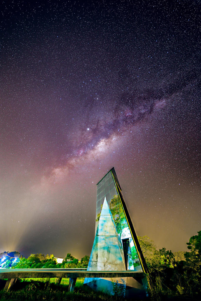 Fotografia vertical e colorida da via láctea e do Obelisco da UFSM. O Obelisco é espelhado e está de perfil. A Via Láctea tem tons de roxo, preto, e amarelo esverdeado mais próximo à superfície.