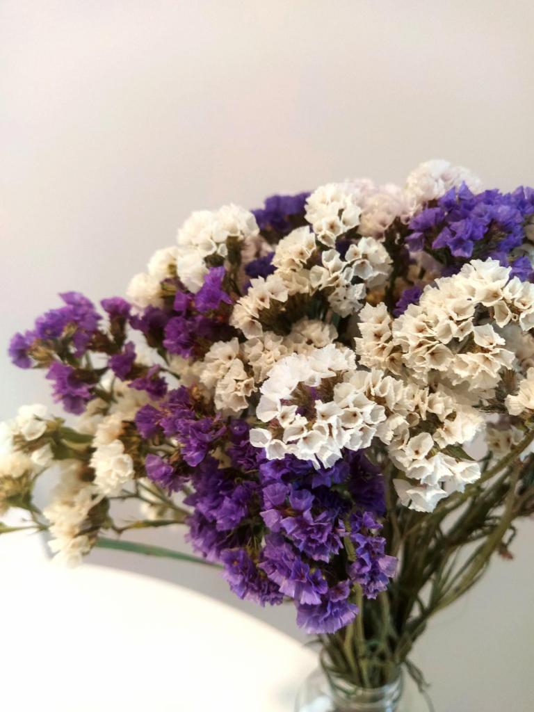 Fotografia vertical e colorida de um buquê de statices roxas e brancas. O buquê é cheio e arredondado. As flores são pequenas e formam círculos. O fundo é uma parede branca.