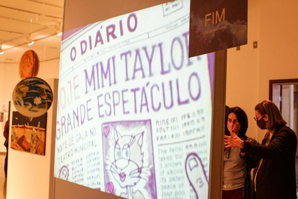 Fotografia horizontal e colorida de uma projeção de slides com recorte de página do jornal "O Diário". Ao fundo, duas mulheres conversam em frente a uma parede branca.