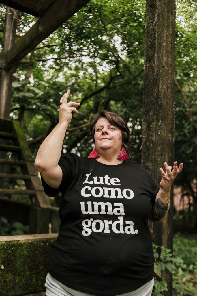 Fotografia horizontal e colorida de uma mulher gorda de pele branca. Ela tem rosto redondo, olhos escuros, cabelos curtos, castanho claros e lisos, com uma franja no lado direito. Tem uma verruga na bochecha esquerda. Usa brincos triangulares vermelhos e grandes. Veste camiseta preta, com a frase em letras grandes e na cor branca, no centro: "Lute como uma gorda." Está com a mão direita levantada, na qual usa um anel preto, e com a mão esquerda na altura do peito. No braço esquerdo, tem uma tatuagem grande. Ela está séria. Ao fundo, em desfoque, paisagem de árvores verdes. Atrás dela, poste de construção antiga e escadas.