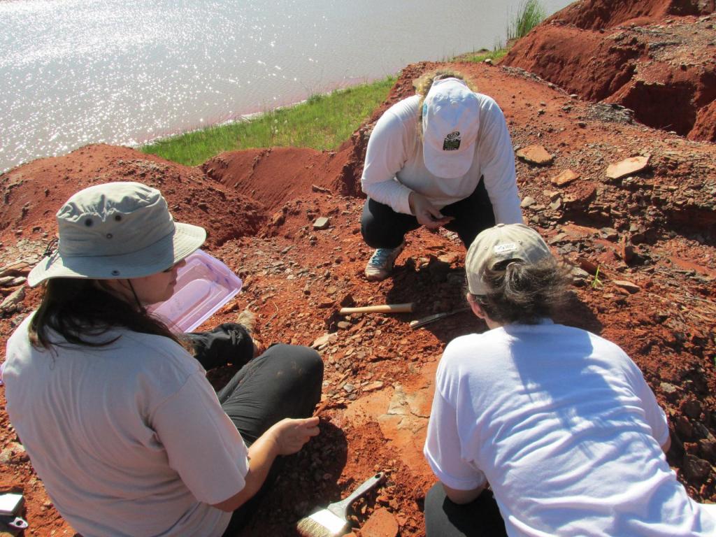 Fotografia horizontal e colorida de três mulheres agachadas sobre solo de terra vermelha. Elas vestem camiseta branca e calça preta, e chapéu de tecido bege ou boné branco. Mexem no solo com ferramentas como martelo, espátula e pincel de limpeza. Ao lado, uma bacia de plástico transparente. Ao fundo, é possível ver água.