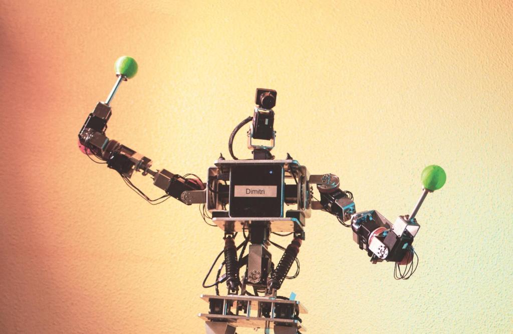 Image Description: Horizontal color photo of a gray robot. In the center of the robot, the name “Dimitri”. It has its arms open and, in each hand, there is a control with a green ball on top. The background is an orange wall.