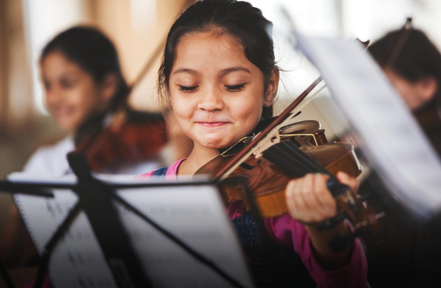 Image Description: A horizontal color photo of a seven-year-old girl playing the violin. She is smiling shyly in front of a black sheet music stand.