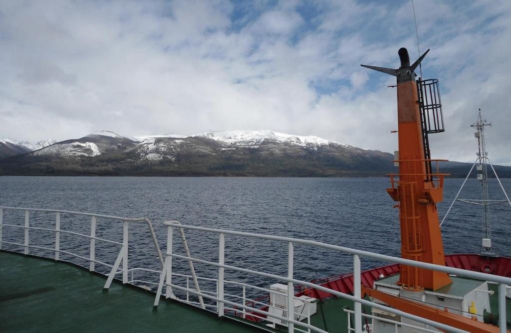 Image Description: Horizontal color photo of a ship. The floor is dark green and the railing is white. On the right-hand side, an orange platform. To the front, a dark blue sea. In the background, snow-capped mountains and blue sky full of white clouds.