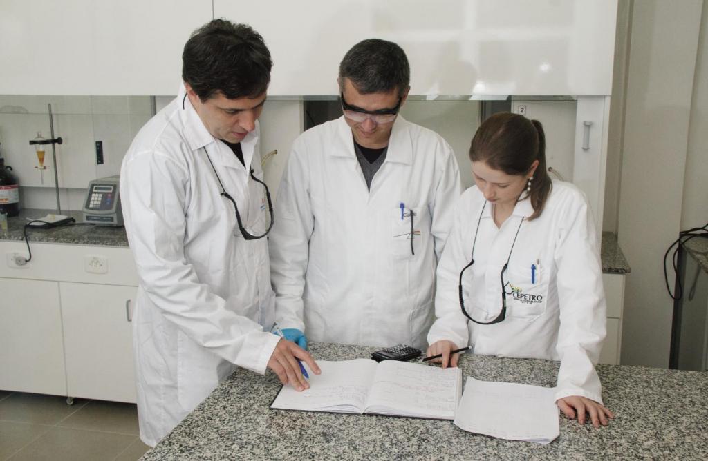 Image Description: Horizontal color photo of three researchers around an open notebook on a marble table. There are two men and one woman. They are wearing white lab coats and have black safety glasses hanging from their necks. In the background, white shelves.