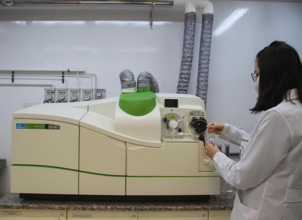 Descrição da imagem: fotografia horizontal e colorida de uma mulher de jaleco branco ao lado de um equipamento multiusuário branco, grande e alongado. Ela está de perfil esquerdo, em primeiro plano; tem pele branca, cabelos castanho escuros, lisos e na altura do ombro; veste jaleco branco com bolso e logomarca da UFSM; usa marcará cirúrgica branca, óculos de grau com armação fina e um anel. Olha para baixo, e segura, nas mãos, um tubo de ensaio abaixo de um cano de plástico fino, que sai do equipamento branco. O equipamento é alongado, grande, em um formato que lembra uma impressora, e tem detalhes em verde vivo, e canoa na cor prata que estão ligados a uma estrutura na parte superior. O equipamento está sobre mesa com tampo de mármore texturizado e cinza. O fundo é uma parede branca.