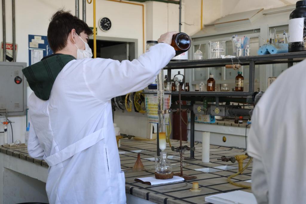 Descrição da imagem: fotografia horizontal e colorida de um homem de pele branca em pé em um laboratório. Ele está em plano geral, e a fotografia foi tirada em um ângulo lateral traseiro. Ele tem pele branca, cabelos castanho escuros e curtos; veste jaleco branco sobre moletom verde escuro e usa máscara descartável branca. Está com o braço direito levantado e segura uma garrafa marrom que despeja um líquido transparente em um tubo de ensaio, preso por um suporte de metal sobre um balão de ensaio. Os frascos estão sobre uma mesa retangular comprida e grande, quadriculada e na cor cinza, em que há vários elementos de laboratório. No centro da mesa, estrutura de metal em formato de prateleira com ingredientes em frascos de vidro e equipamentos gerais. No canto direito, detalhe de jaleco branco. No fundo, porta aberta e armário cinza.