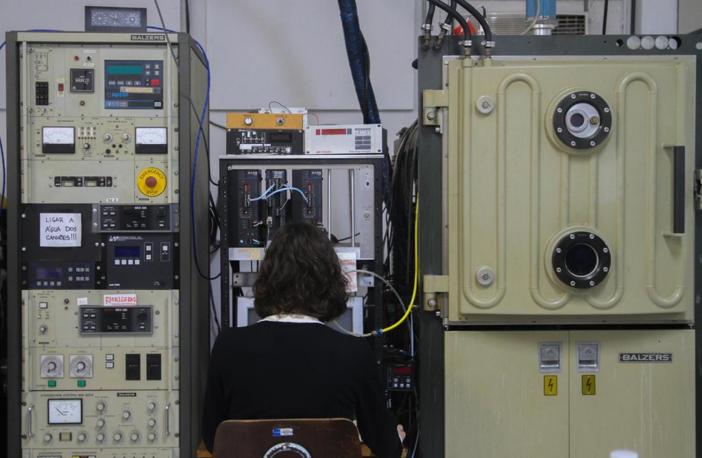 Descrição da imagem: fotografia horizontal e colorida de uma mulher, de costas, sentada em frente a um computador antigo e em meio a estruturas verticais de equipamentos na cor bege. A mulher tem cabelos castanho escuros, levemente ondulados e na altura do ombro; veste suéter preto sobre gola de camisa branca. A cadeira é de madeira, do tipo escolar. O computador é antigo e cinza. Acima dele, equipamentos escuros com botões, fios e controles coloridos. De cada lado dela, há um equipamento bege. Do lado esquerdo da imagem, o equipamento está cheio de botões e visores coloridos. Do lado direito da imagem, o equipamento tem dois círculos pretos na parte superior, e dois miolos de chave na parte inferior. O fundos é uma parede branca.