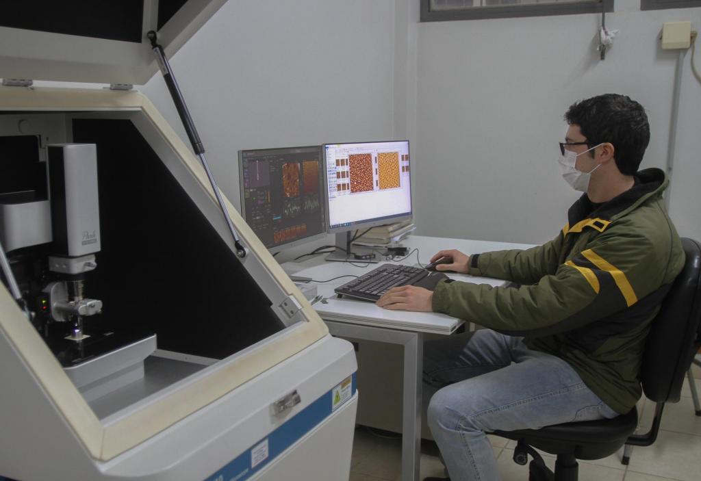 Descrição da imagem: fotografia horizontal e colorida de um homem de pele branca sentado em frente a dois monitores de computador. Ele está de perfil esquerdo, tem pele branca, cabelos castanho escuros e curtos; veste jaqueta verde militar com listras amarelas e pretas e calça jeans clara; usa máscara descartável branca e óculos de grau com armação preta. Está com a mão sobre um mouse preto, que fica ao lado de um teclado preto e em frente a dois monitores de computador com programas de análise de dados abertos. O fundo é uma parede branca.