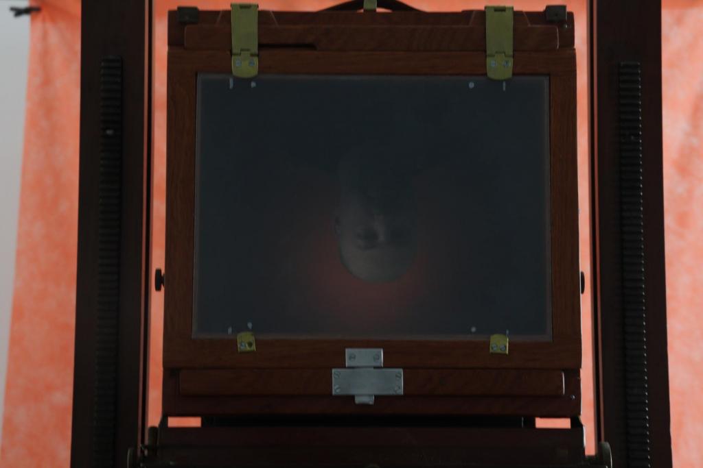 Descrição da imagem: Fotografia horizontal e colorida do visor da câmera. Nele, a imagem de um homem aparece de ponta cabeça. O visor tem moldura de madeira escura. No fundo, tecido laranja.