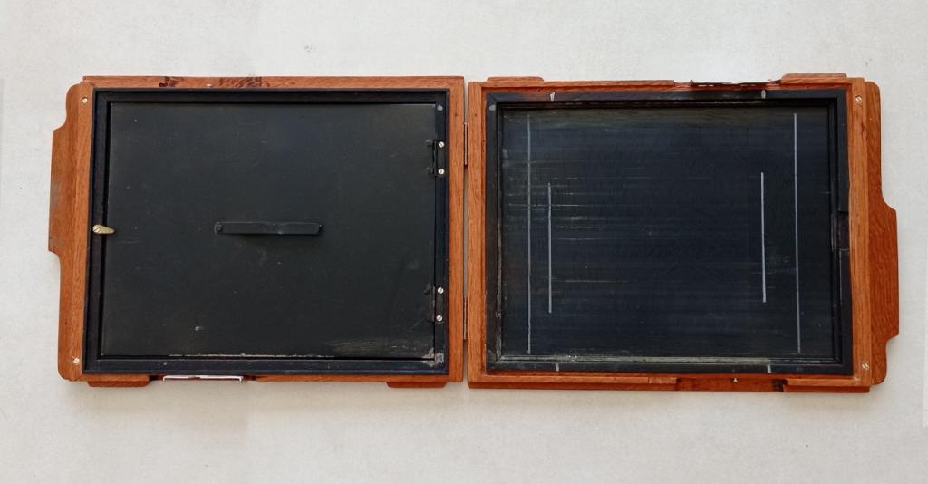 Descrição da imagem: Placa de madeira pequena e retangular aberta. As bordas são de madeira em tom de marrom caramelo, e a parte interna é preta. O fundo é branco.