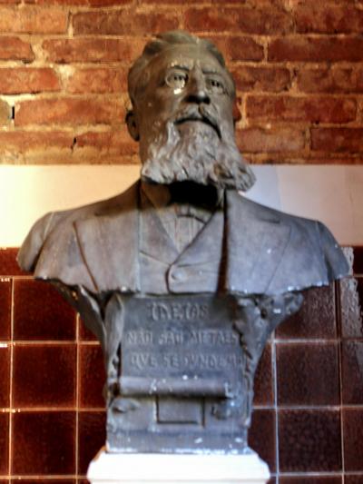 Descrição da imagem: Fotografia vertical e colorida do busto de Gaspar Silveira Martins, em tom de chumbo, sobre base branca. O fundo é uma parede de tijolos a vista.