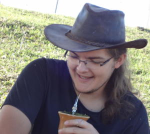 Foto de homem cabelos longos castanhos tomando chimarrão e usando chapéu preto e óculos de grau
