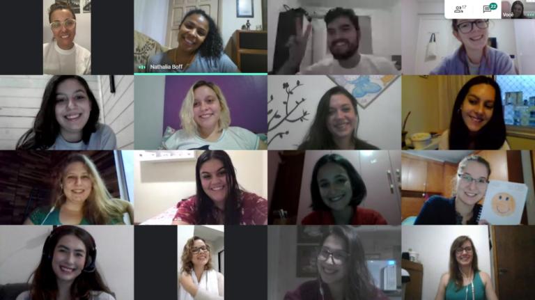 Captura de tela retangular e colorida de uma videoconferência. 16 pessoas aparecem na imagem, dispostas em quatro fileiras; o conjunto forma um mosaico. Na fila superior, duas mulheres de pele branca, um homem de pele branca e uma mulher de pele negra. Na segunda fileira, quatro mulheres de pele branca. Na terceira fileira, quatro mulheres de pele branca. Todos sorriem. Quatro mulheres possuem cabelo loiro claro, quatro possuem cabelo loiro escuro e sete possuem cabelo castanho. O homem tem cabelo e barba castanho escuros e faz um sinal de “v” com a mão direita.