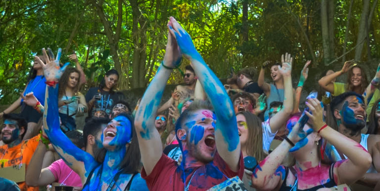 Descrição de Imagem: Fotografia horizontal e colorida, tirada na diagonal, da esquerda para a direita, de estudantes ao ar livre. Em primeiro plano, está um menino e uma menina, ambos de braços levantados em movimento e pintados com tinta azul nos braços e rosto. A menina aparece à esquerda da imagem, sua pele é branca e seu cabelo é liso, castanho na altura dos ombros; ela está com os olhos fechados e a boca aberta. O menino está no centro da imagem, sua pele é branca e tem cabelo loiro; ele veste uma camiseta vermelha; suas mãos juntam-se no alto, acima de sua cabeça. Cerca de 15 pessoas estão atrás dos dois estudantes centralizados, dispostas de maneira aleatória, todas pintadas de tinta de cores azul, vermelho, verde e laranja, com a cor azul predominante. Atrás destes, estão 8 estudantes de pé, estes não estão pintadas. A maioria das pessoas apresentam expressão facial de comemoração. Na parte superior ao fundo, árvores em tons de verde.