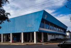 Descrição de Imagem: Fotografia horizontal e colorida da diagonal de um prédio de dois andares na cor azul. Embaixo, no primeiro andar, nove pilares de sustentação na cor branca; a fachada é azul e, na parte inferior direita da fachada, está escrito "UNIÃO UNIVERSITÁRIA”, em caixa alta, letras grandes e na cor branca. No lado direito do prédio, há janelas espelhadas e “31” na ponta, em cor branca. Na parte superior, o céu em azul com nuvens brancas. No chão, pedras em paralelepípedos na cor cinza. Um carro preto em movimento se direciona para a rua localizada ao lado direito do prédio.