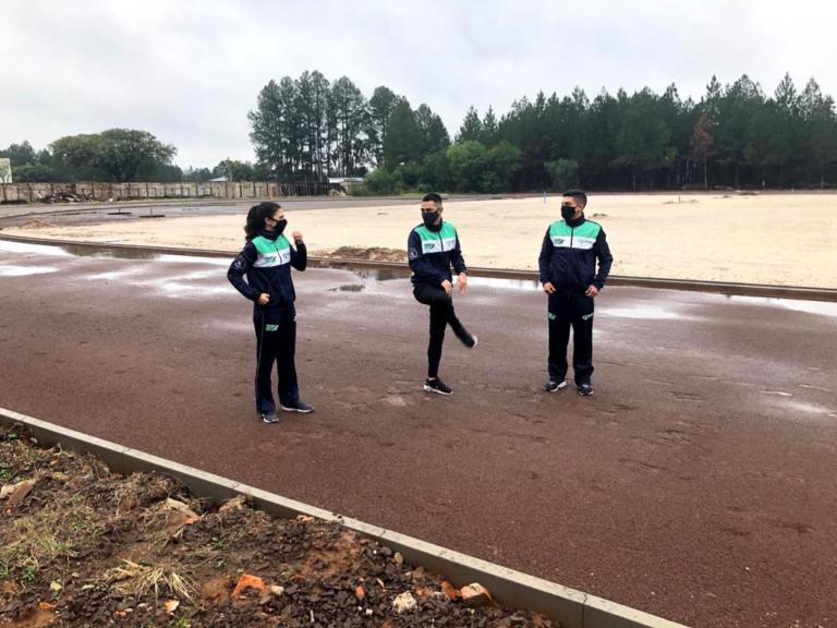 Foto horizontal de três atletas aquecendo, lado a lado, na pista de atletismo: uma mulher branca de cabelo longo preto e dois homens brancos de cabelo curto. Ao fundo, uma grande área arborizada.