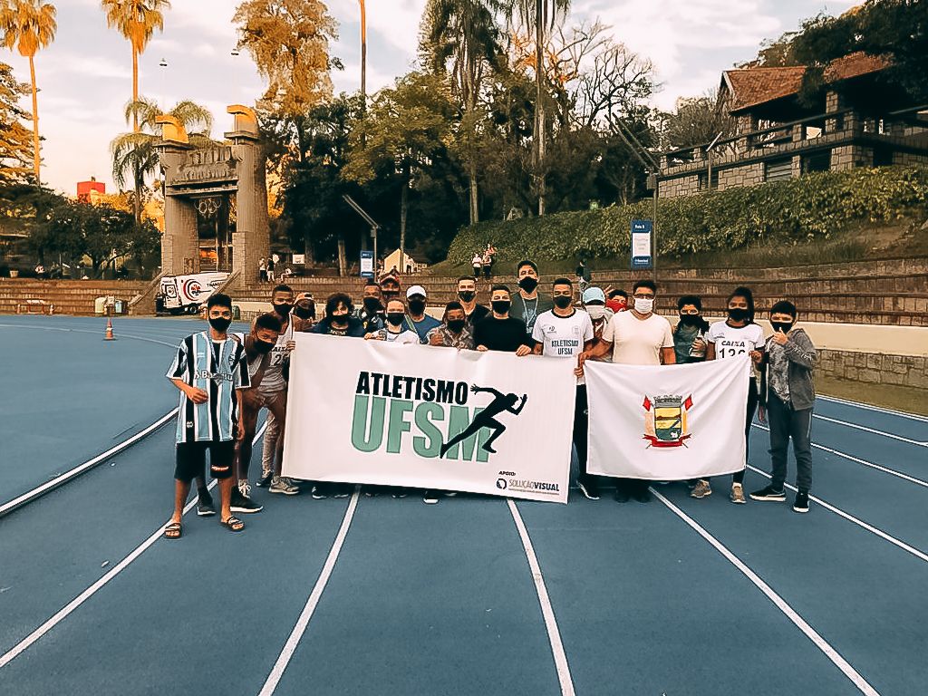 Foto horizontal com os atletas abraçados, todos eles usando máscaras, em uma pista de atletismo. Eles seguram duas faixas, uma grande à esquerda e outra menor, à direita. Na faixa da esquerda, a escrita "Atletismo UFSM”. Na faixa da direita, a bandeira de Santa Maria. Ao fundo, a arquibancada e uma área arborizada.