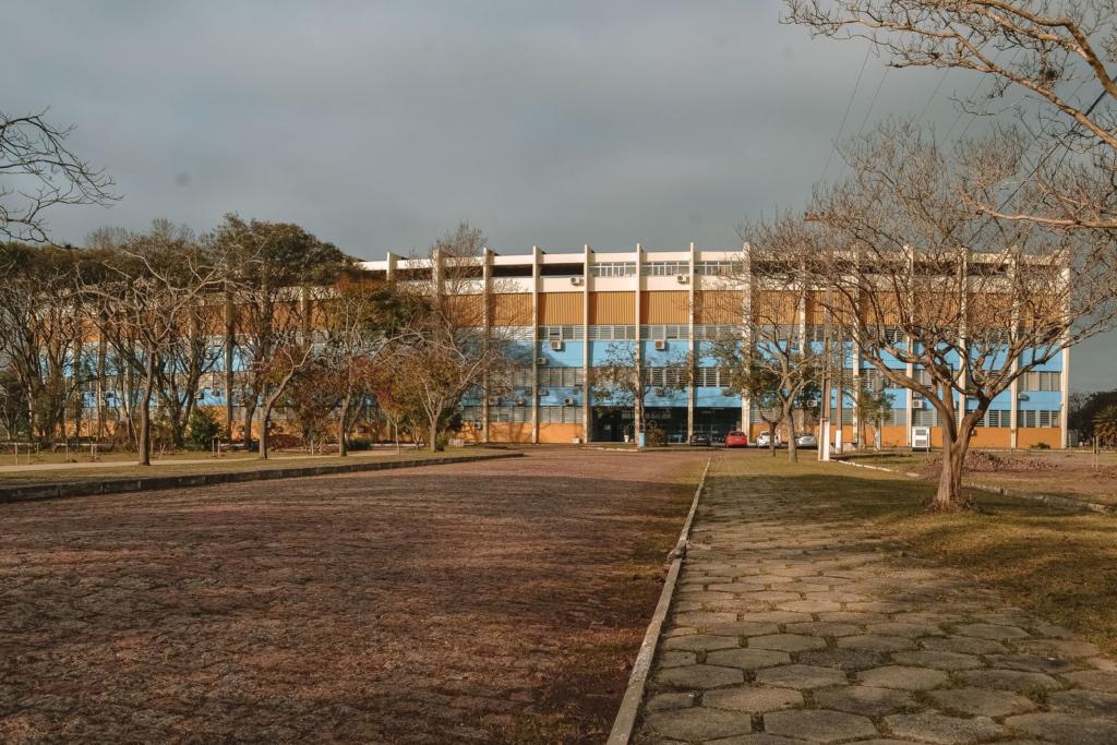 Foto horizontal da vista frontal de um prédio de cinco pavimentos. O prédio está pintado nas cores azul, amarelo e branco. Algumas árvores estão em frente ao prédio e o céu está nublado.