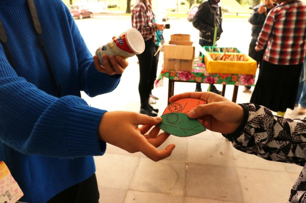 Foto horizontal; predominantemente laranja; registro de uma troca no Escambo Solidário; pessoa dos ombros para baixo usando um blusão azul e calça preta segurando uma lata de achocolatado em uma mão e realizando a entrega de Ecopilas com a outra. Pessoa com casaco preto e branco recebendo a moeda; no fundo mesa de pescaria e pessoas circulando no Hall do Restaurante Universitário