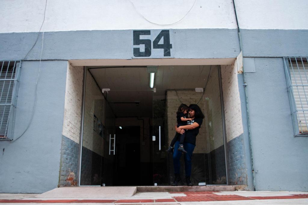 Fotografia horizontal e colorida com uma mulher e uma criança abraçadas na entrada de um prédio da Casa do Estudante Universitária. A mulher é negra, de pele clara, tem cabelos escuros, lisos e curtos; está com o rosto inclinado para baixo e os olhos fechados; veste camiseta preta com letras brancas, calça jeans com lavagem escura e calçado preto. Ela segura em seu colo uma menina de faixa etária próxima a três anos. A menina é negra, de pele clara, tem cabelos escuros, levemente ondulados e na altura do ombro; com a cabeça inclinada para baixo e os olhos fechados; veste camiseta, calça e calçado pretos. A entrada do prédio é azul acinzentado; as paredes internas são brancas na parte superior, e há uma porta de vidro. O lado esquerdo da porta está aberto e o outro fechado. Nos extremos laterais da imagem há partes de duas janelas e grades azuis. Na parte inferior há uma calçada de concreto cinza com detalhes de piso tátil avermelhado e uma rampa de acesso com leve inclinação.