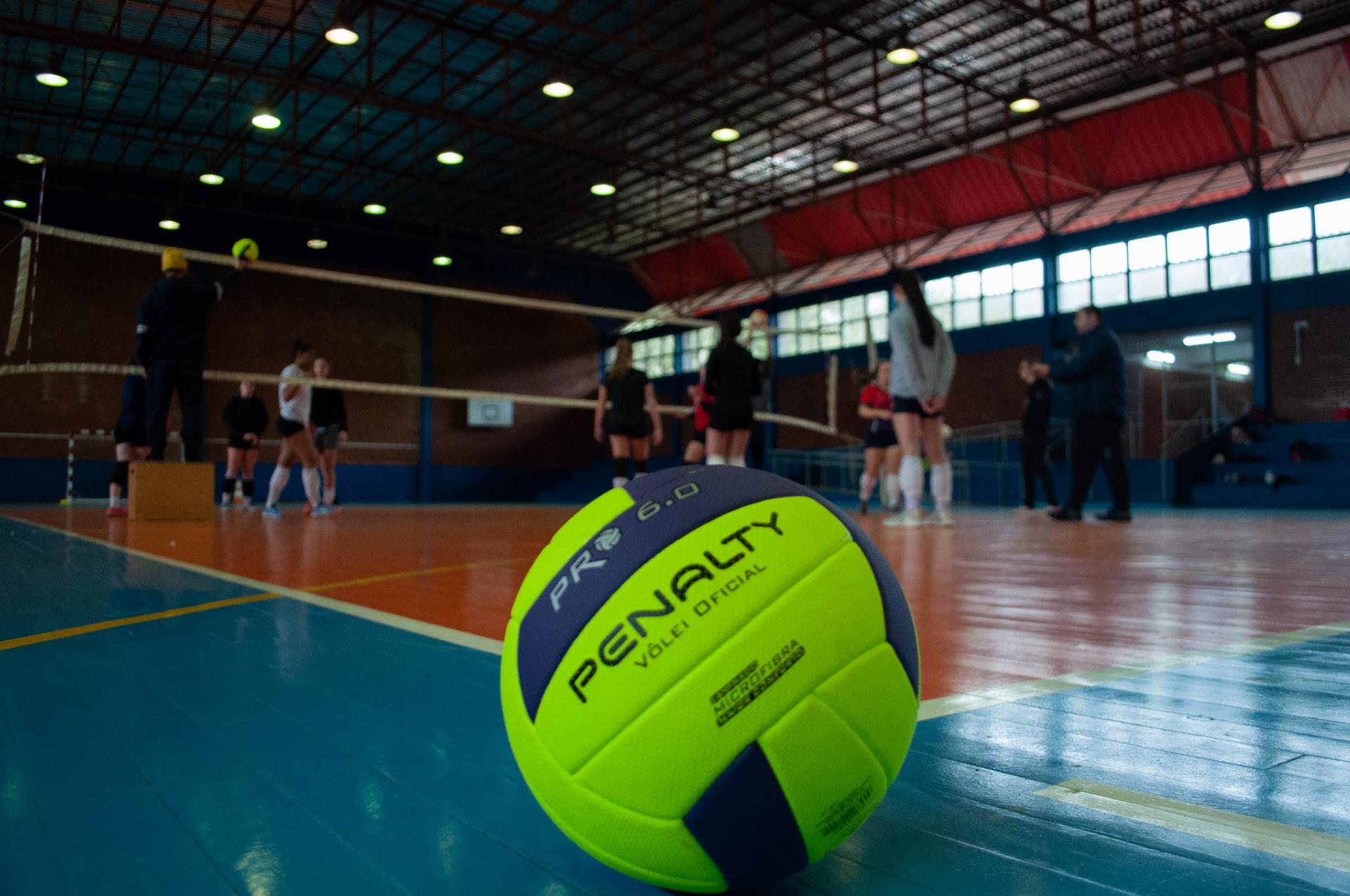 Foto em sentido horizontal, colorida, com uma bola de voleibol em destaque, dentro de uma quadra esportiva. A bola tem cores amarelo e azul. Ao fundo, com uma imagem desfocada, está a quadra de vôlei na cor laranja e as extremidades na cor azul, a rede de vôlei transparente com bordas brancas e oito atletas, todas mulheres que estão em pé, dos dois lados da quadra. Na lateral esquerda da imagem, um homem, com roupas pretas e uma touca amarela, em cima de um caixote marrom e segura uma bola de vôlei próxima a rede. Ao fundo o teto com estrutura de metal escura e iluminação em 19 lâmpadas circulares.