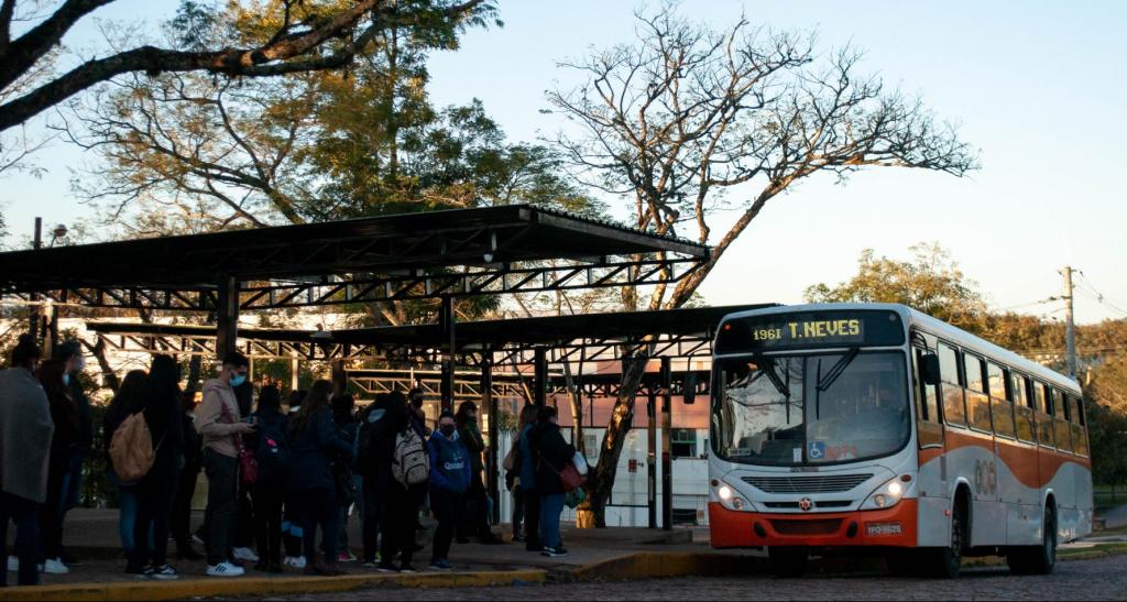  Foto horizontal colorida de um ponto de ônibus com várias pessoas e um ônibus em tons de laranja e branco. A imagem está em um plano geral fechado e o ônibus está levemente na diagonal direita. A parte frontal do ônibus é branca, e tem um letreiro luminoso com o texto “T. Neves” e o número “1961” ao lado; o parabrisa tem um ícone de acessibilidade para pessoas usuárias de cadeira de rodas nas cores azul e branca, e o parachoque é da cor laranja. A lateral é dividida horizontalmente na cor laranja e branca por uma linha ondulada, em que a parte branca ocupa o espaço inferior. No lado esquerdo, há várias pessoas paradas embaixo de uma estrutura metálica na cor preta, em formato de toldo. A maioria está de costas ou de perfil, algumas seguram mochilas e bolsas. As pessoas que se encontram de lado ou de frente usam máscara. O fundo tem  árvores altas e com copa com poucas folhas, o céu na cor azul claro, sem nuvens, e a fachada de um prédio. 