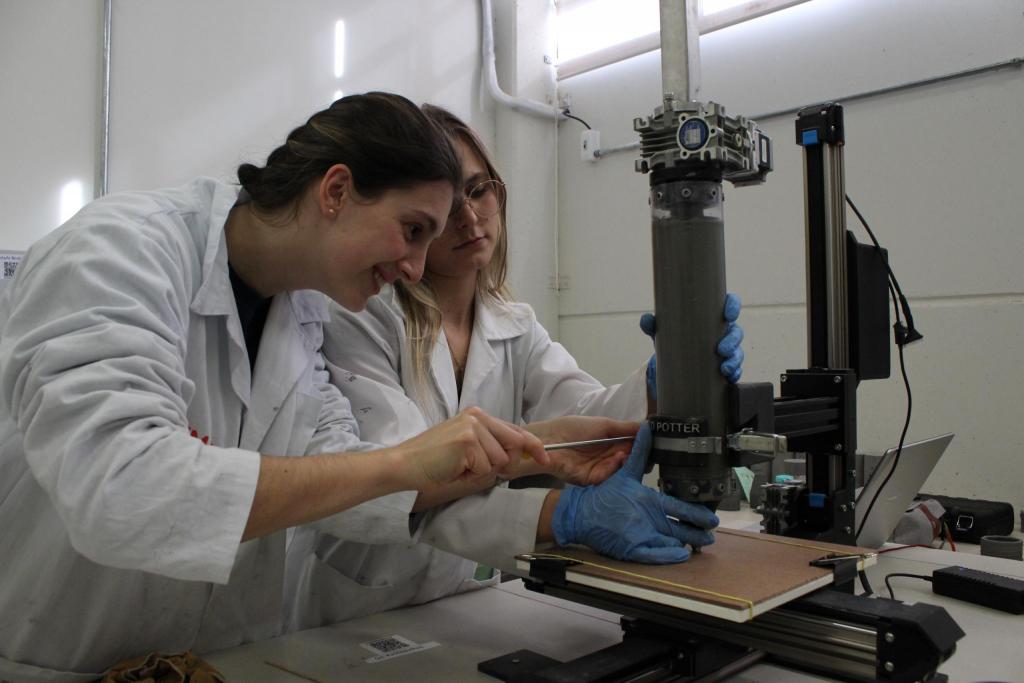 Fotografia horizontal, colorida, em plano médio de duas mulheres no laboratório. A mulher, em primeiro plano, está de perfil, é branca, com uma expressão de felicidade, tem cabelos castanhos, presos numa trança. Veste um jaleco branco e usa brincos de pérolas brancas. Nas mãos, uma ferramenta não identificada na cor cinza, encosta em um tubo cinza escuro de uma impressora 3D. Ao lado dela,  outra mulher, em segundo plano. Ela está de frente, é branca, tem os cabelos loiros claros. Veste um jaleco branco e  usa luvas de plástico azul e óculos de grau na cor dourada. As mãos estão encostadas no tubo da impressora. A impressora é da cor preta, tem elementos metálicos e está sobre uma mesa branca. No fundo da imagem, um notebook na cor prata. As paredes do laboratório são brancas. 
